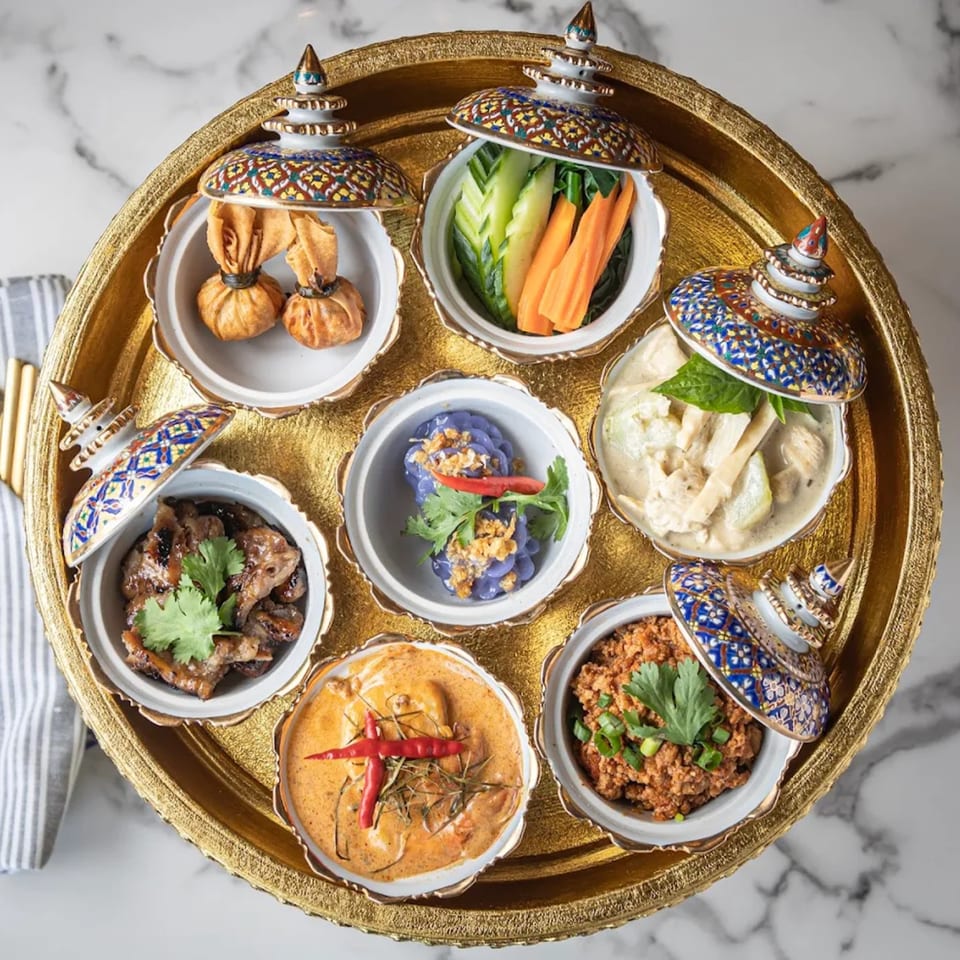 A golden tray holds seven small, ornate bowls of various Thai dishes, including curries and vegetables.
