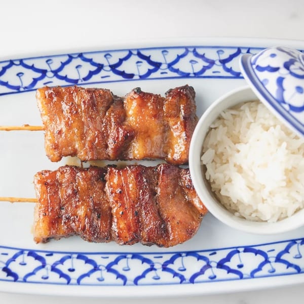 Two grilled pork skewers and a small bowl of white rice on a white and blue patterned plate.