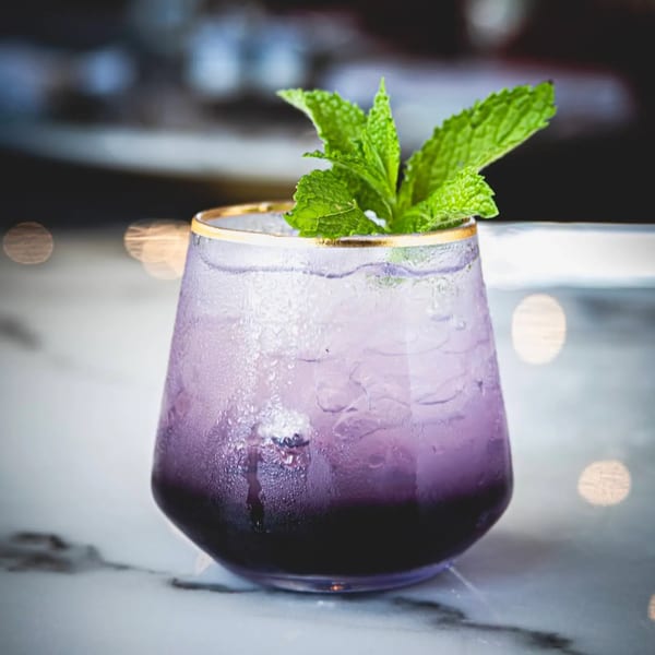 A vibrant purple cocktail with ice and a mint garnish, served in a gold-rimmed glass.