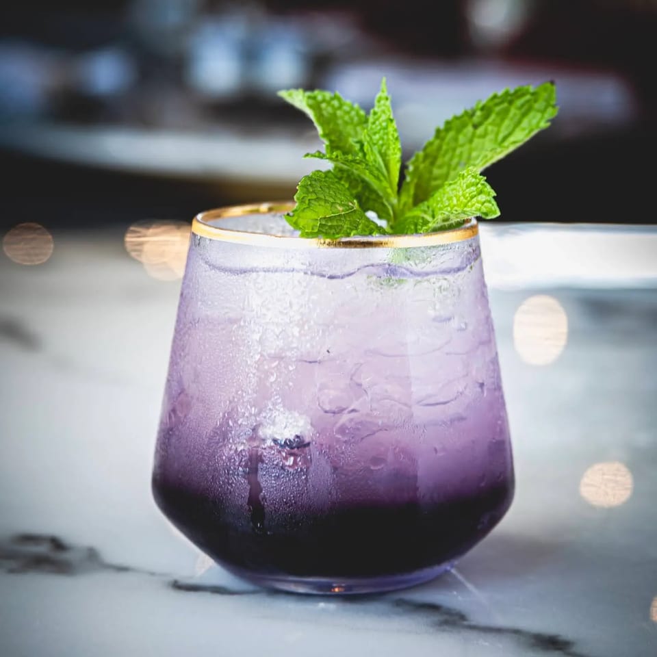 A vibrant purple cocktail with ice and a mint garnish, served in a gold-rimmed glass.