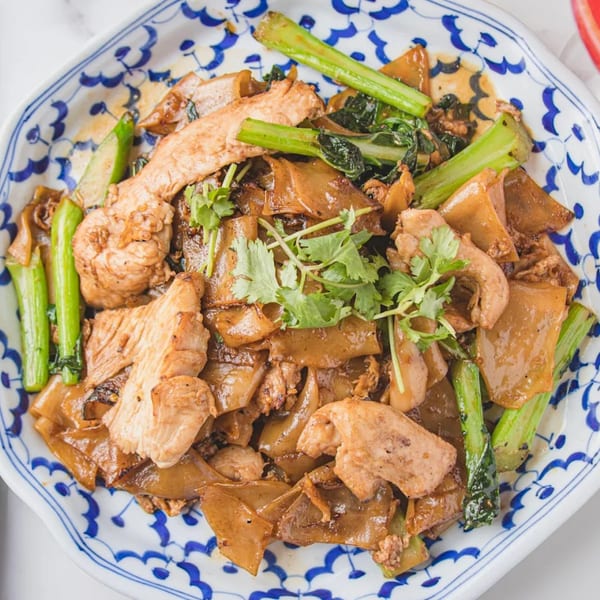 Close-up of Pad See Ew with wide rice noodles, chicken, Chinese broccoli, and cilantro on a blue and white plate.