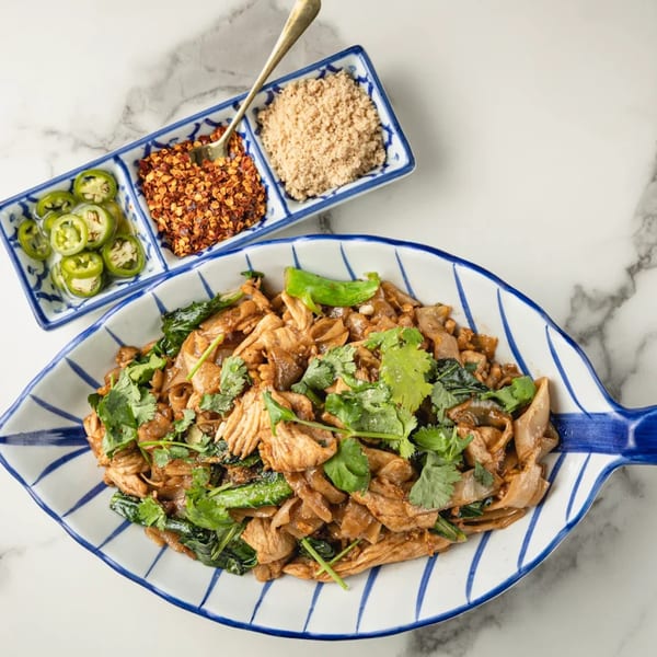 Thai stir-fried noodles with chicken, greens, and cilantro, served with chili flakes and sliced jalapeños.