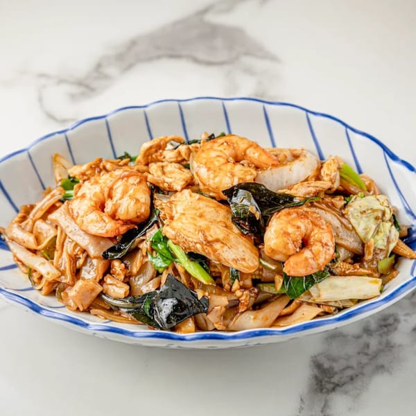 Close-up of Pad See Ew with shrimp, chicken, wide noodles, and green vegetables in a blue-striped bowl.