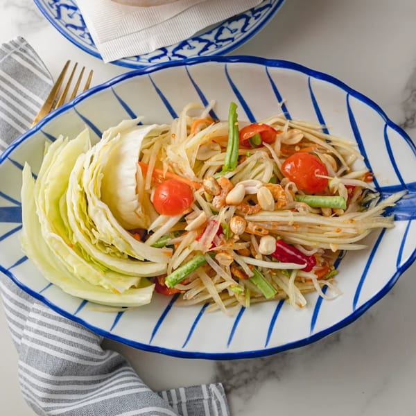 Som Tum, a Thai papaya salad with peanuts, tomatoes, and cabbage, served on a blue and white plate.