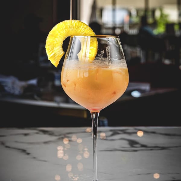 A vibrant orange cocktail in a wine glass, garnished with a pineapple slice, on a marble table.